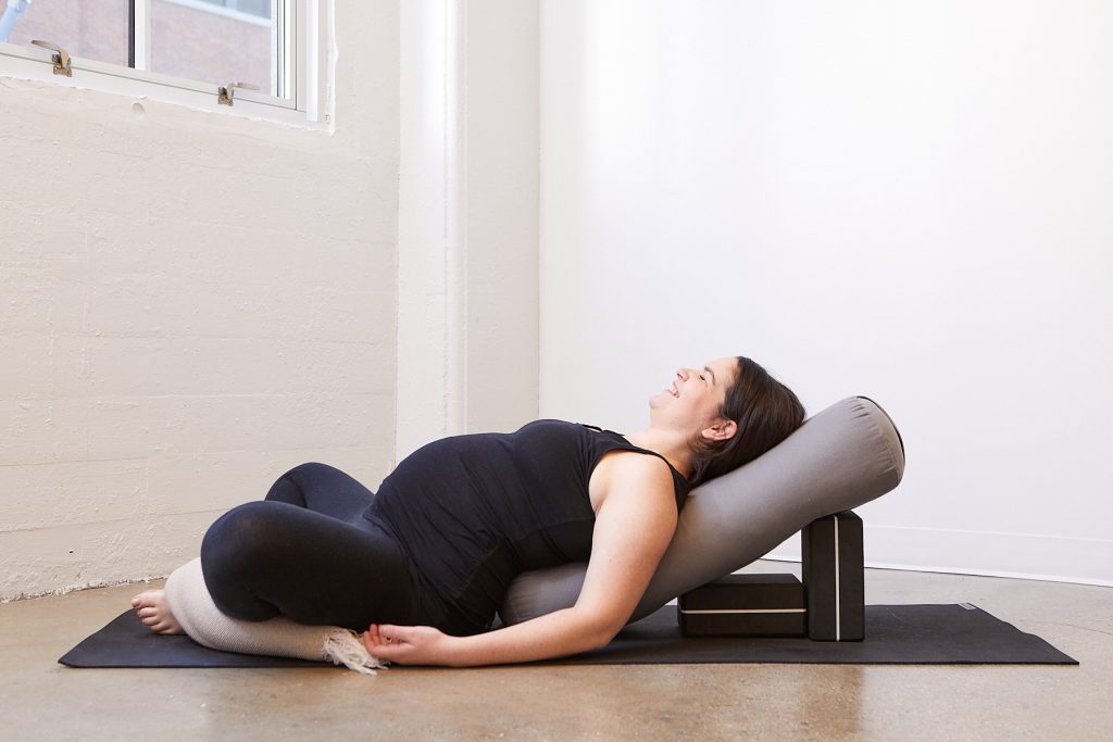 Prenatal yoga pose of a pregnant woman in all black in supta baddha konasana with a blanket rolled to support her legs