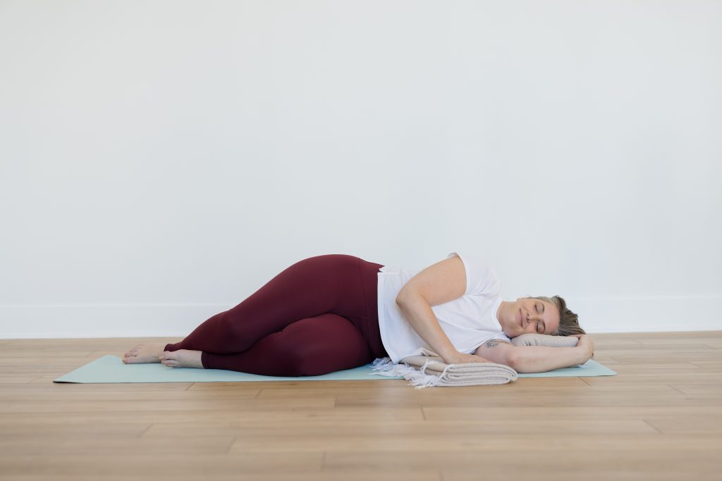A woman in deep maroon yoga pants lying on her side in a restorative yoga pose with her eyes closed on a light blue yoga mat.