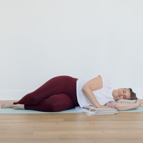 A woman in deep maroon yoga pants lying on her side in a restorative yoga pose with her eyes closed on a light blue yoga mat.