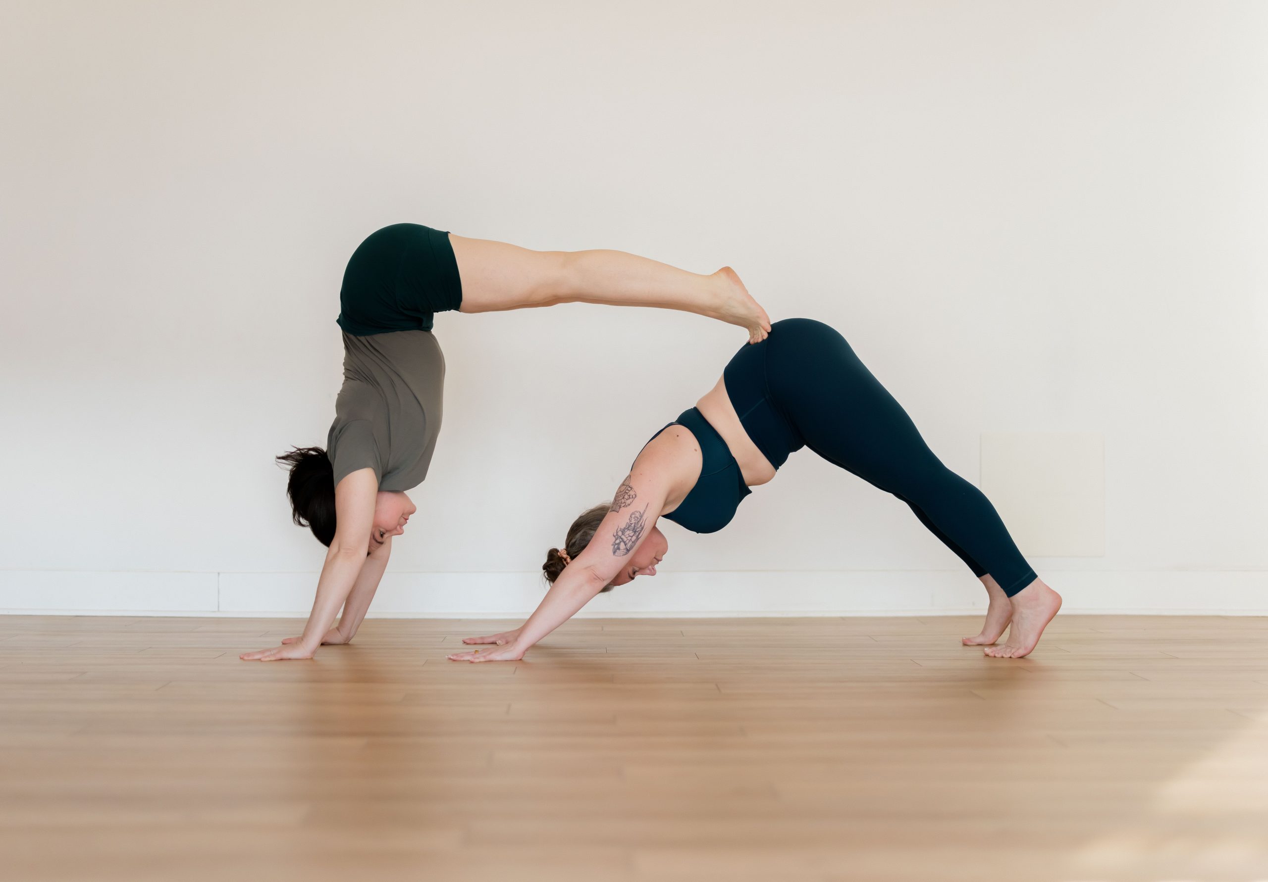 Yogi in L-Shape Handstand being supported by their yogi friend in Downward Facing Dog, resting their toes against the sacrum of the yogi in Down Dog