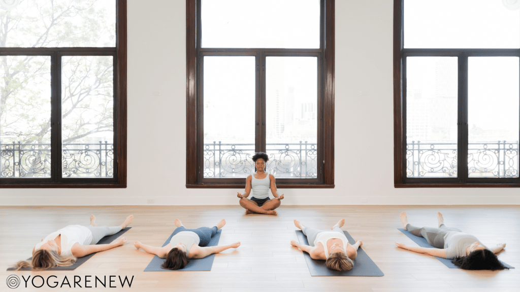 Yoga teacher sitting in front of a class in Sukasana while the students lie down in Savasana