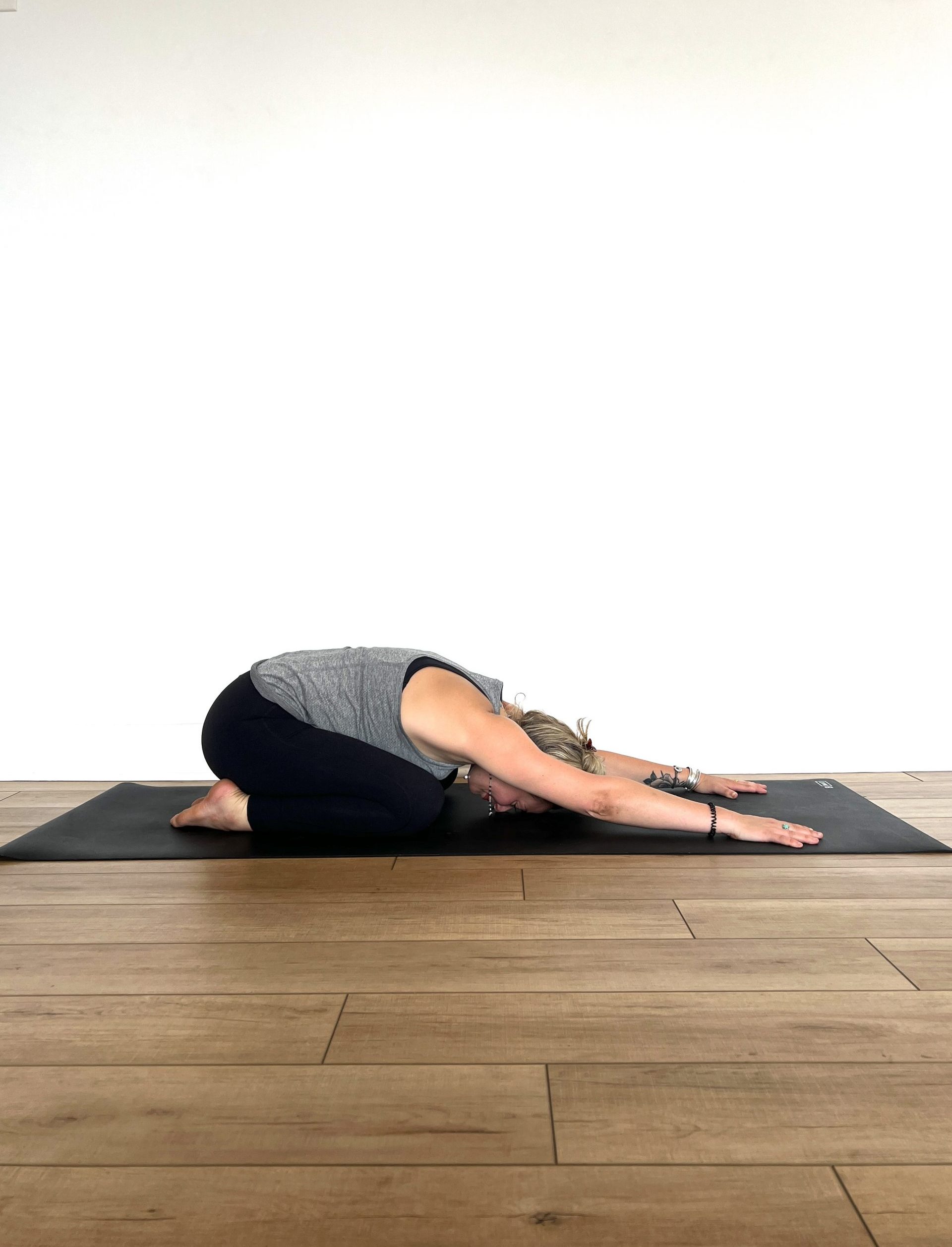 A woman in Balasana (child's pose) in black leggings and a grey tank top on a black yoga mat against a white wall
