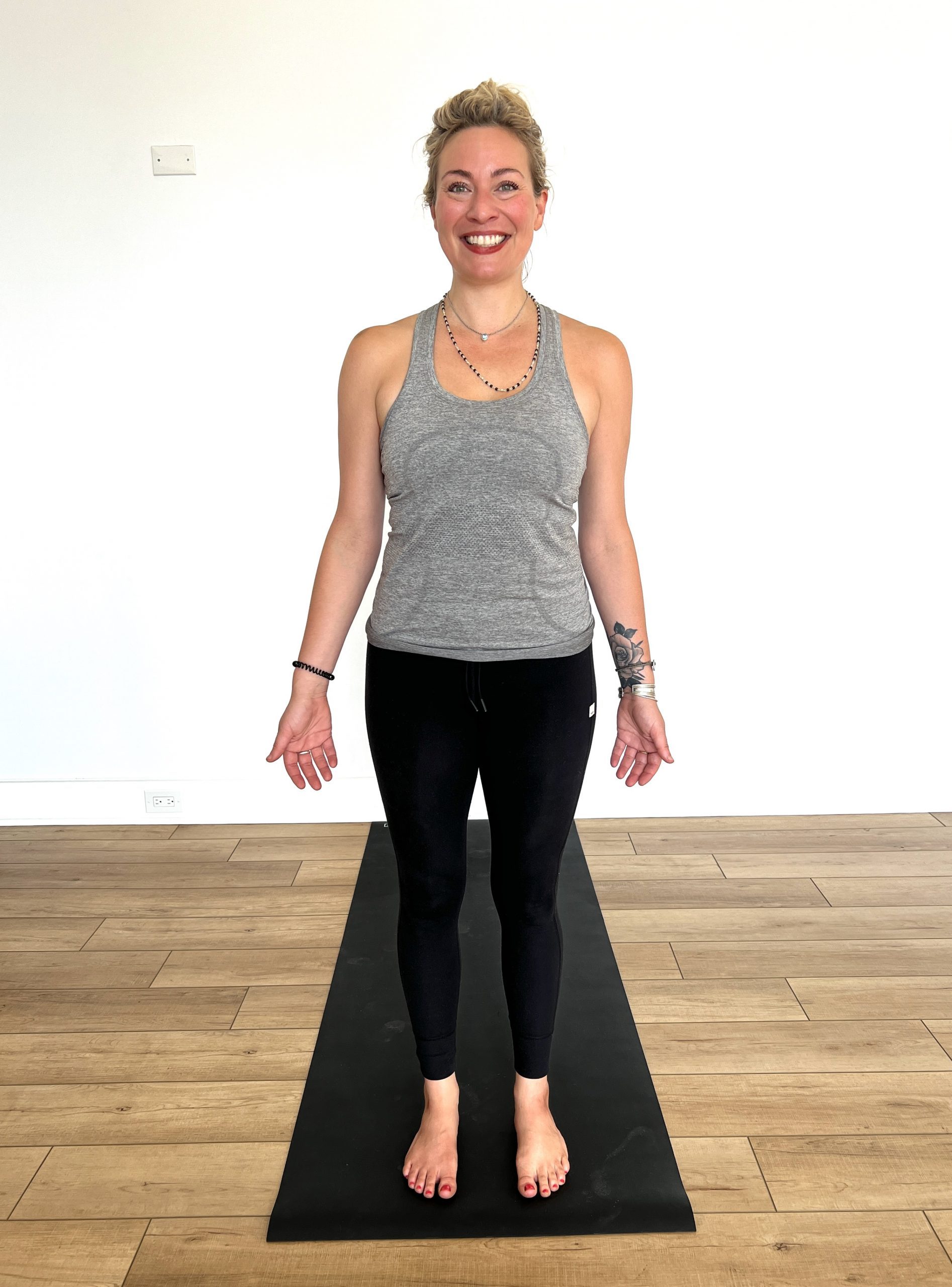 Woman standing in Tadasana (mountain pose) at the top of a black yoga mat in a grey tank top with black leggings, smiling at the camera with palms down beside her facing forward