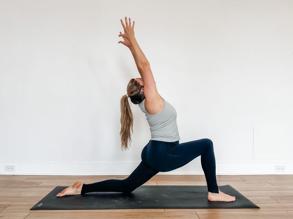 Woman on a black yoga mat and light blue shirt doing crescent lunge with a ponytail and arms up overhead