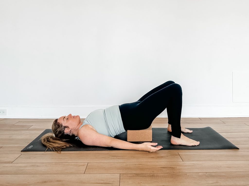 Woman on a black yoga mat with a cork block underneath her sits bones for supported bridge pose