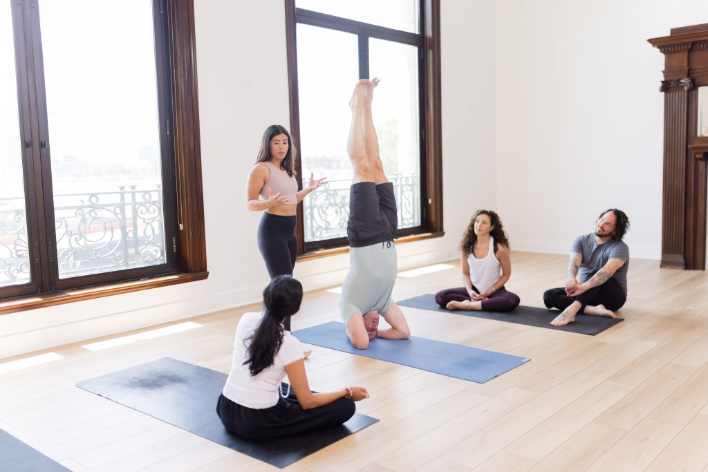 A yoga teacher in the front of the room talking through someone in headstand while 3 other students watch