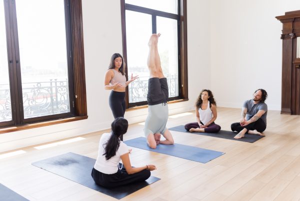 A yoga teacher in the front of the room talking through someone in headstand while 3 other students watch