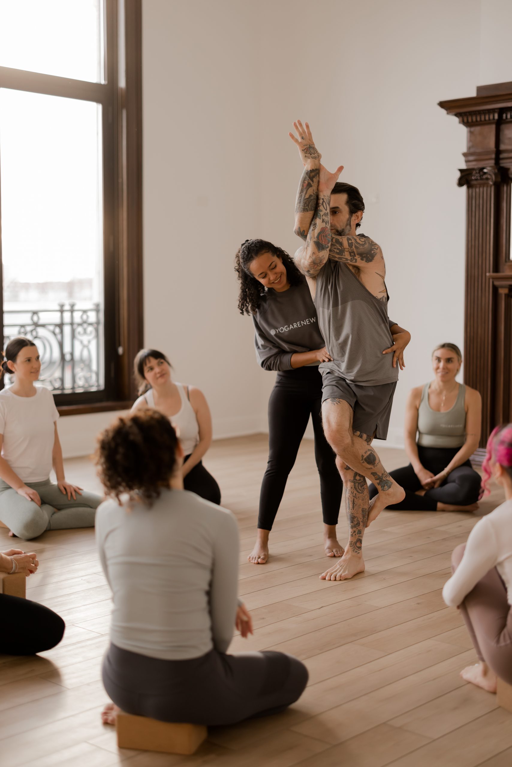 A dark-haired, young female yoga student giving a hands-on assist to the senior yoga teacher, tattooed male, Ronen Kauffman