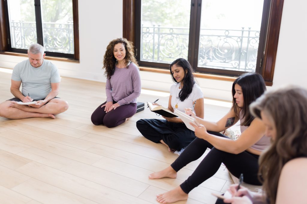 A woman in a purple top and purple leggings subbing a yoga class with students sitting around her taking notes