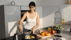 A woman in a white tank-top cooking an Ayurvedic meal on a kitchen counter with grapefruits, tea and a plant in front of her.