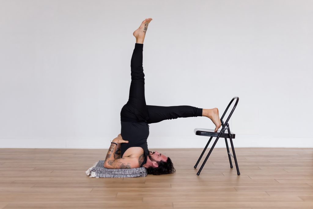 Patrick Franco of YogaRenew in an all black yoga outfit on a black and white blanket kicking up off of a black chair into shoulder stand with his hands supporting his back