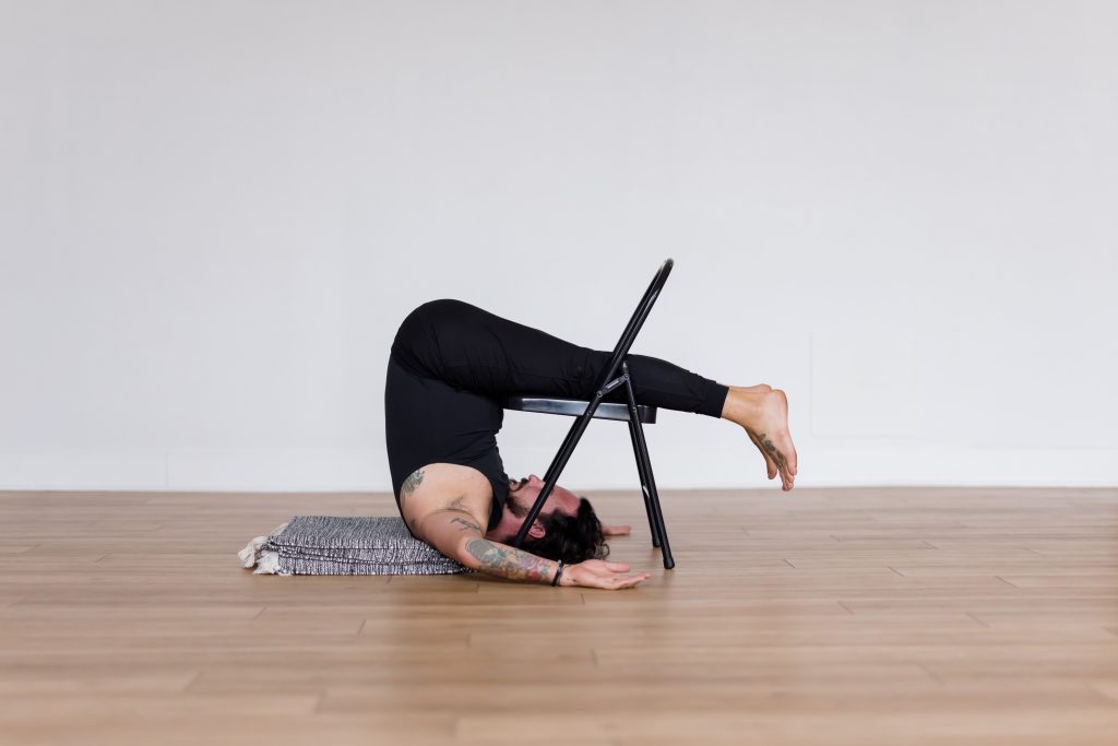Patrick Franco of YogaRenew in an all black yoga outfit with a black and white blanket underneath his shoulders in Halasana (plow pose) over a black yoga chair