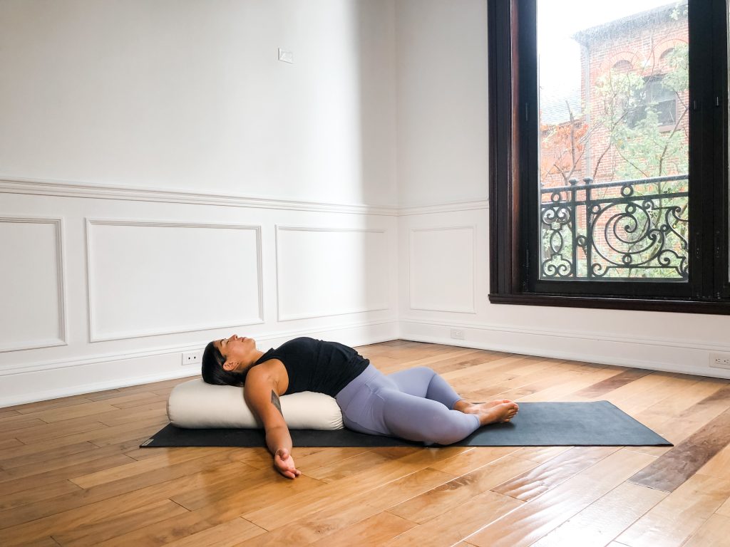 Yogi in purple yoga pants reclining over a linen bolster in Supta Baddha Konasana