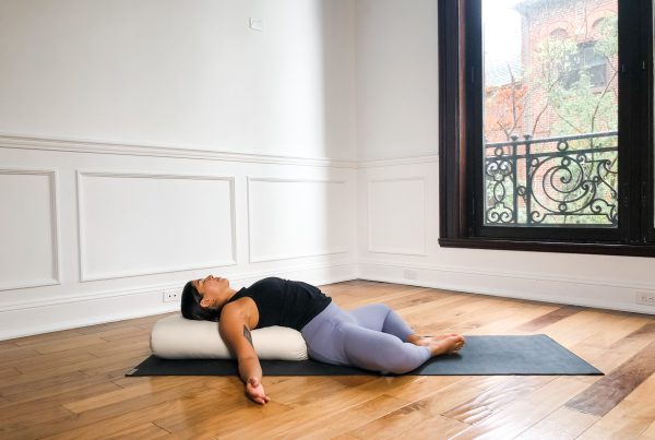 Yogi in purple yoga pants reclining over a linen bolster in Supta Baddha Konasana