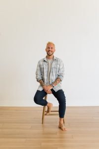 Patrick Franco sitting on a stool in a plaid button down and cuffed jeans with no socks or shoes, smiling at the camera