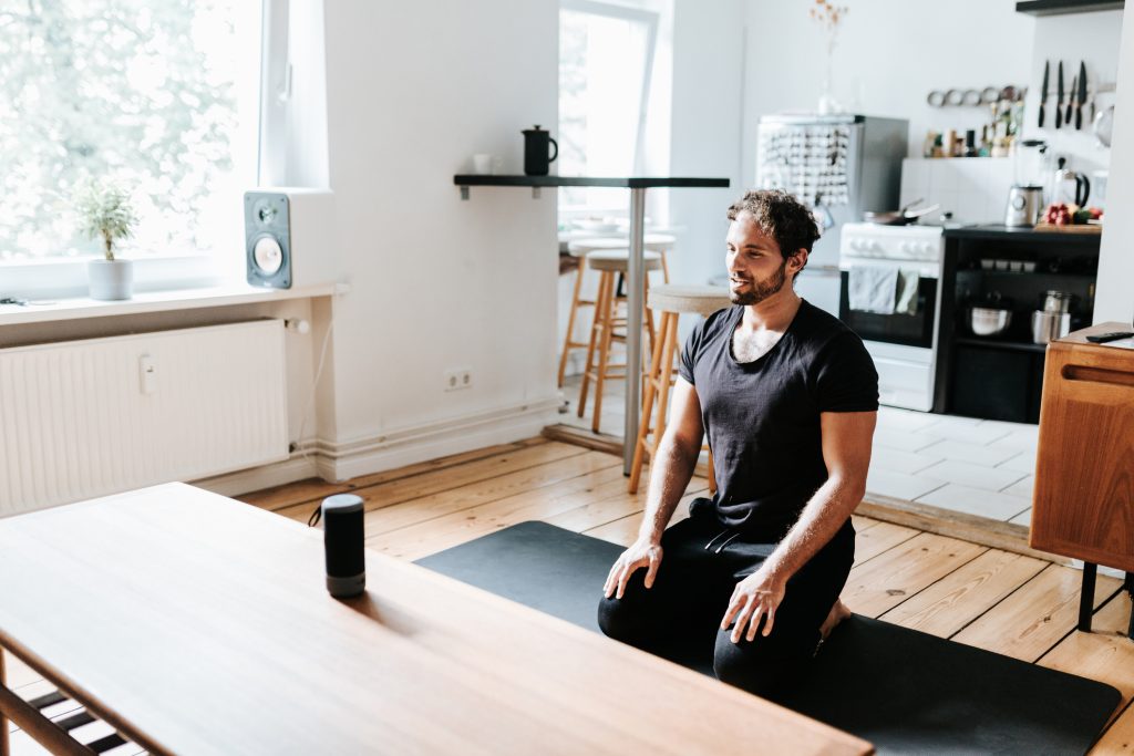 Yogi sitting in Virasana on a yoga mat following instructions from an AI voice assistant like Alexa or Siri from a speaker sitting on a table in front of them