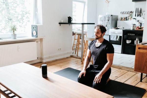 Yogi sitting in Virasana on a yoga mat following instructions from an AI voice assistant like Alexa or Siri from a speaker sitting on a table in front of them