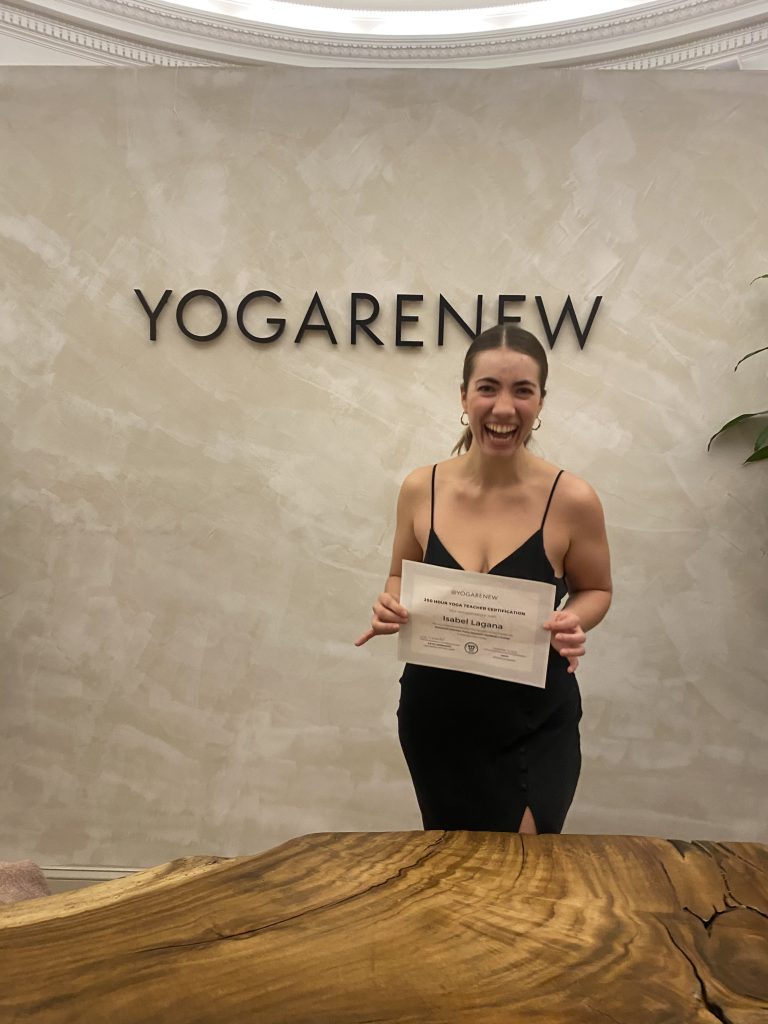 Isabel Lagana in front of the YogaRenew Headquarters front desk holding her 200 hour ytt certificate