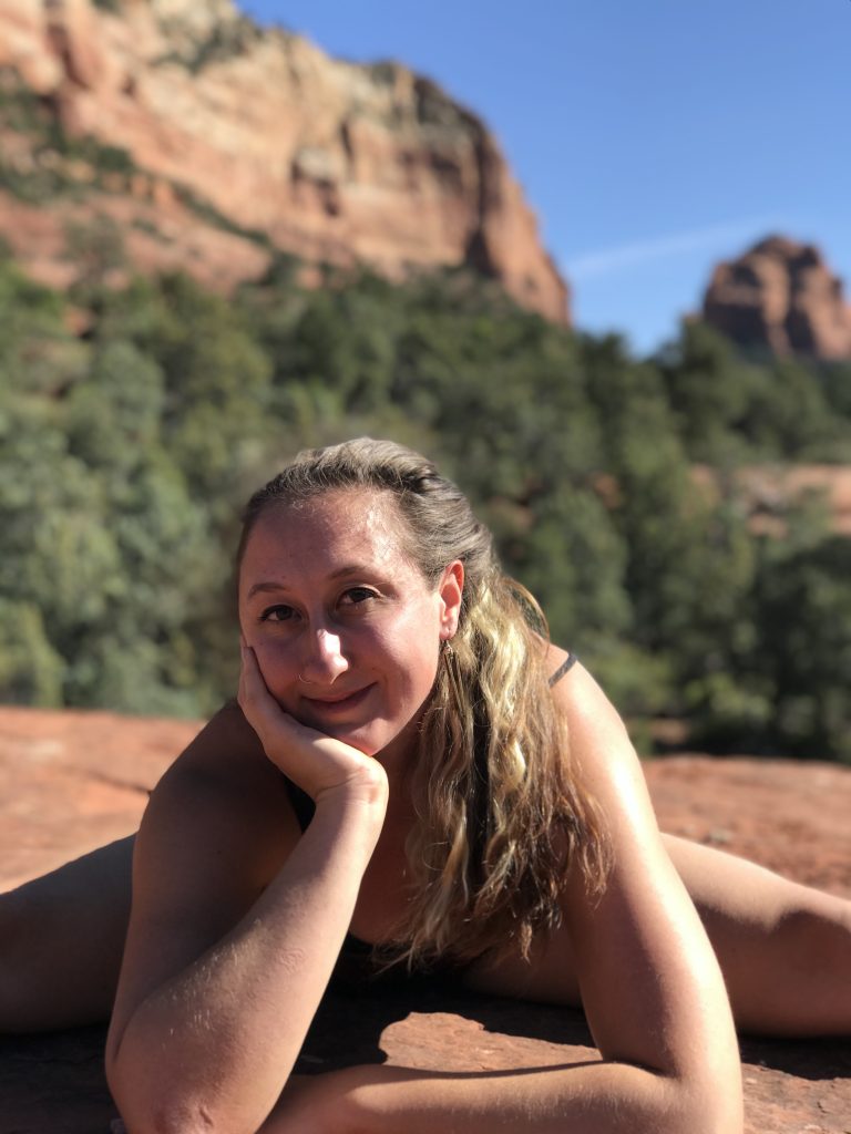 Magda in a seated wide legged forward fold, leaning forward to rest her gently smiling face in her palm.